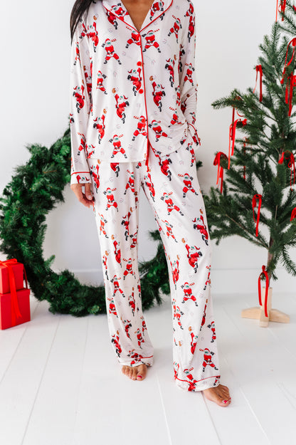 A person wearing a white long-sleeve pajama set with an allover Santa Claus print, standing barefoot in a bright room with a Christmas tree and wreath in the background.