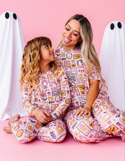 A woman and a young girl wearing matching pink patterned pajamas sit together on a pink floor, with white ghost decorations behind them. The pajamas have a colorful food-themed print with words like 'pizza', 'burger', 'soda', and 'coffee'.