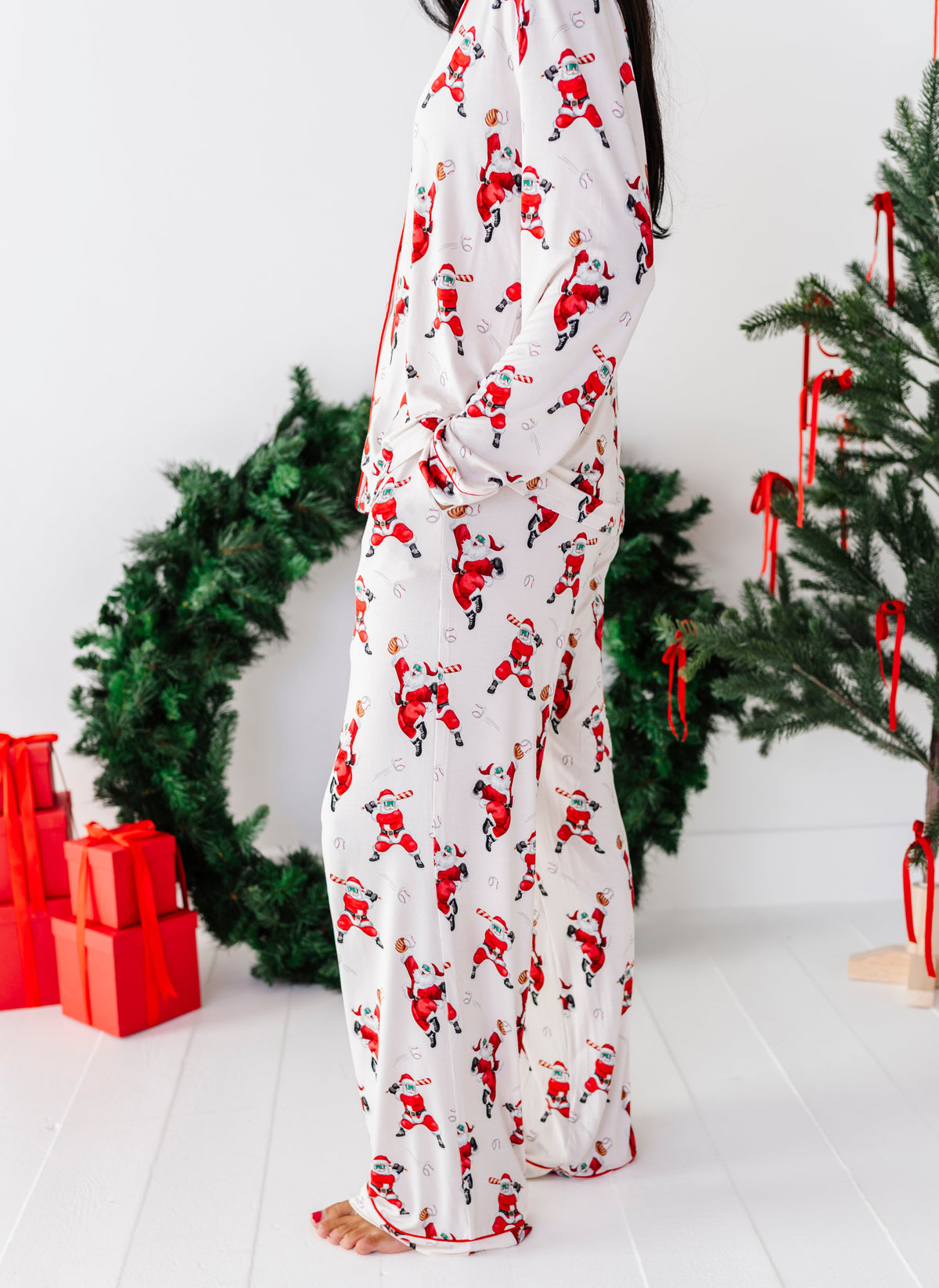 A person wearing a white Santa Claus patterned pajama set stands barefoot in a festive room with a Christmas tree, wreath, and red wrapped gifts.