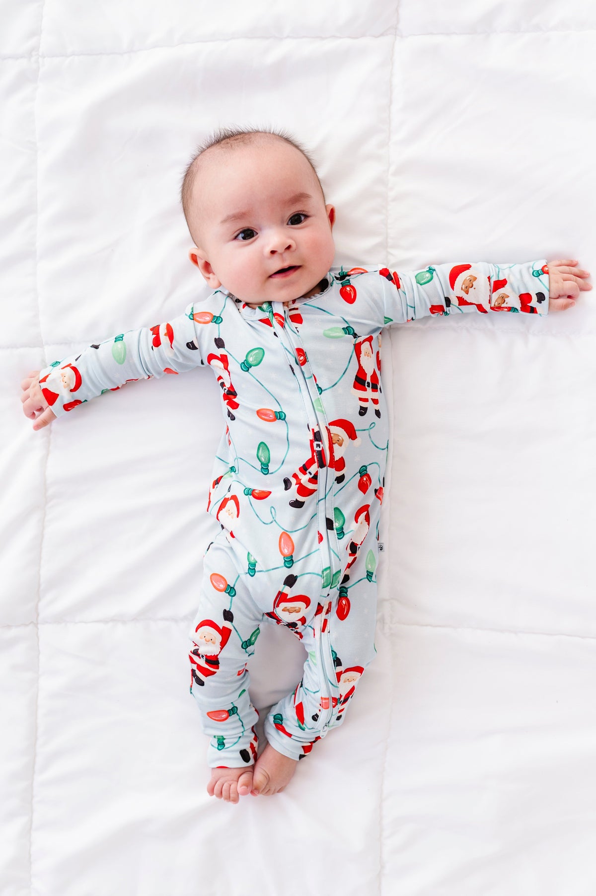 A baby boy wearing a blue bamboo convertible romper featuring Santa Claus hanging from multi-colored Christmas lights