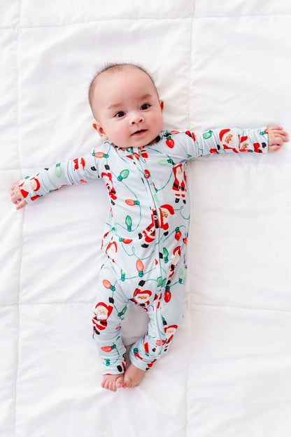 A baby boy wearing a blue bamboo convertible romper featuring Santa Claus hanging from multi-colored Christmas lights