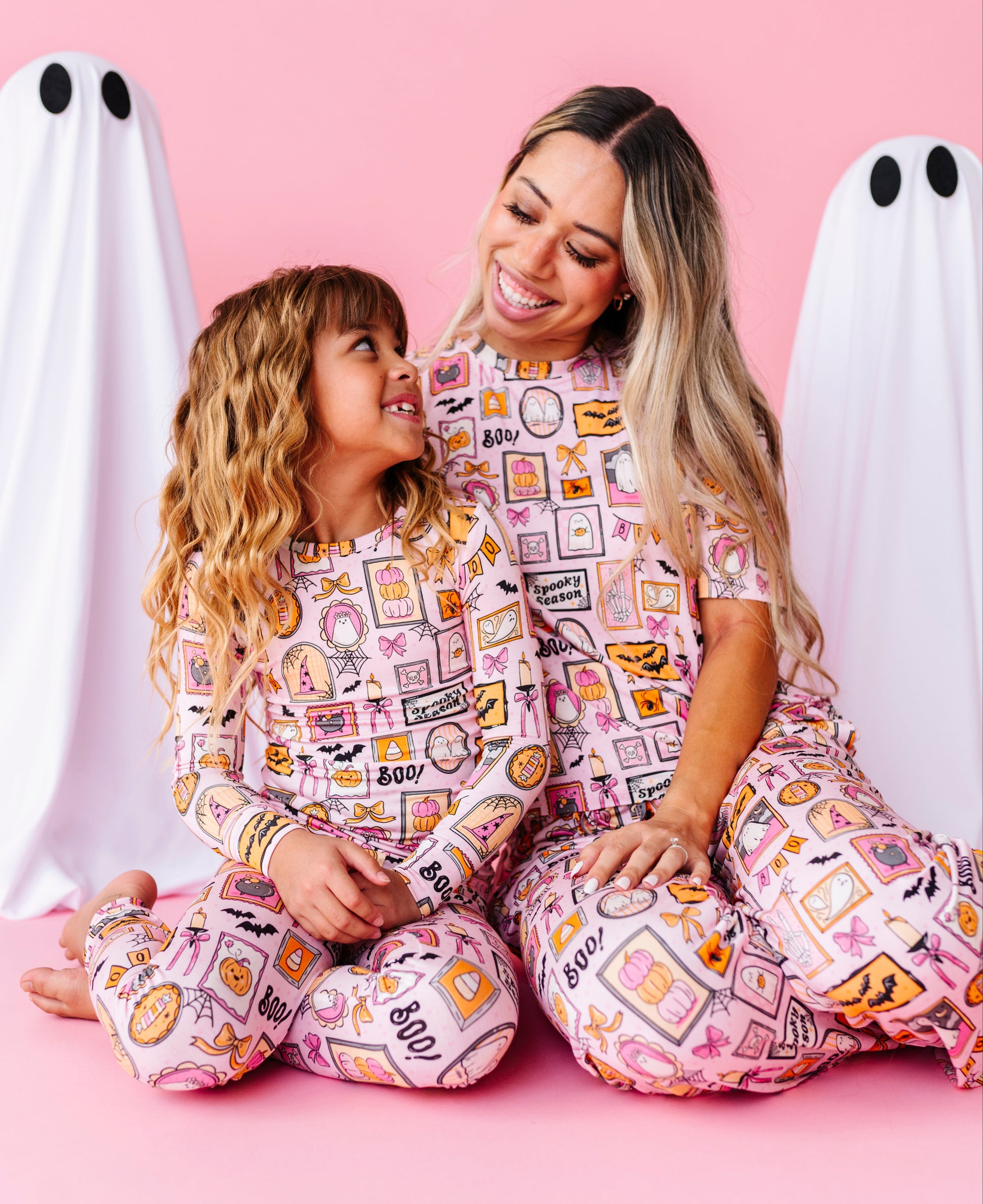 A woman and a young girl wearing matching pink patterned pajamas sit together on a pink studio floor, with white ghost props on both sides. The pajamas have a multicolor print featuring Halloween-themed illustrations and text like 'BOO', 'spooky', and candy graphics.