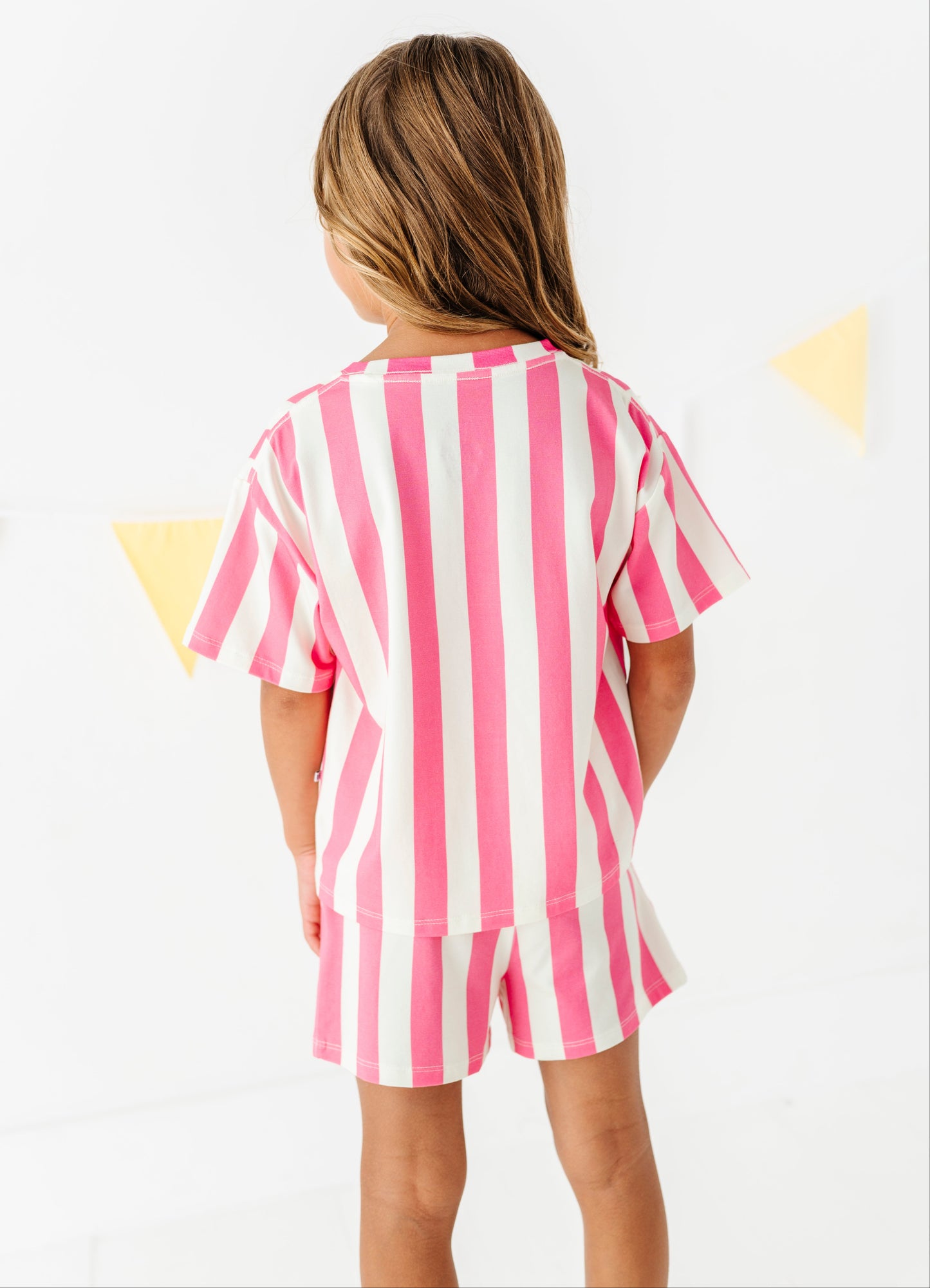 A young girl viewed from behind wearing a pink and white vertically striped short-sleeve t-shirt and matching striped shorts, set against a bright white background with subtle yellow triangular bunting.