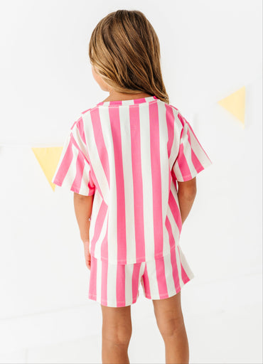 A young girl viewed from behind wearing a pink and white vertically striped short-sleeve t-shirt and matching striped shorts, set against a bright white background with subtle yellow triangular bunting.