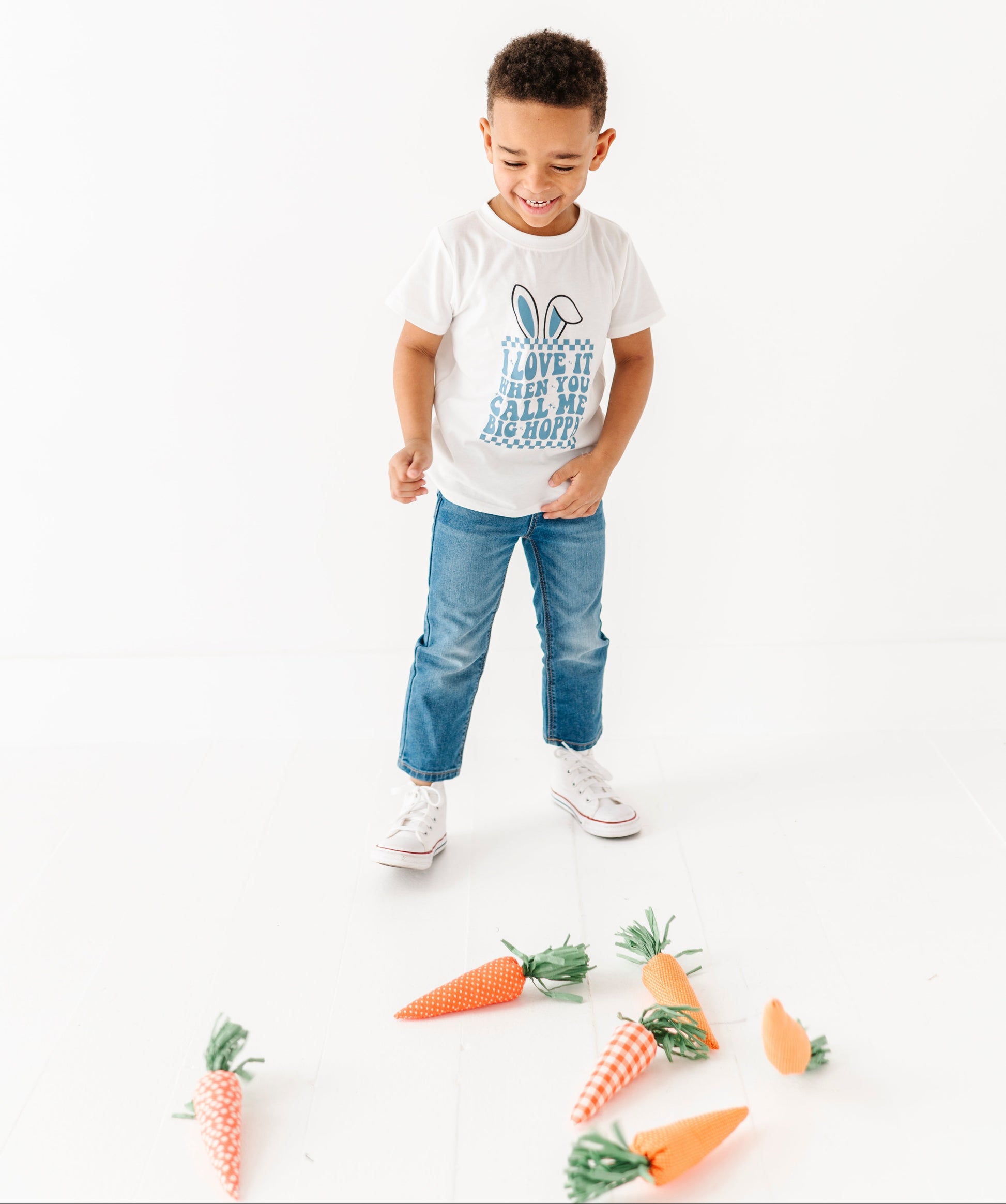 Child wearing a white t-shirt with text and blue jeans, standing on a white background with decorative carrots.