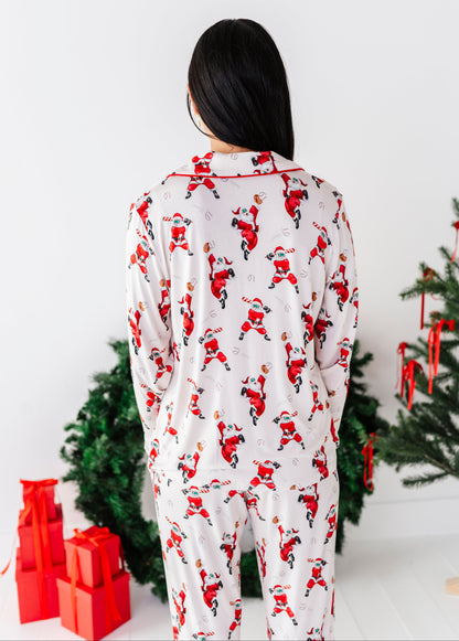 A person viewed from behind wearing a white long-sleeve pajama set with an allover Santa Claus print, standing in front of a white wall with Christmas trees and red wrapped gifts nearby.