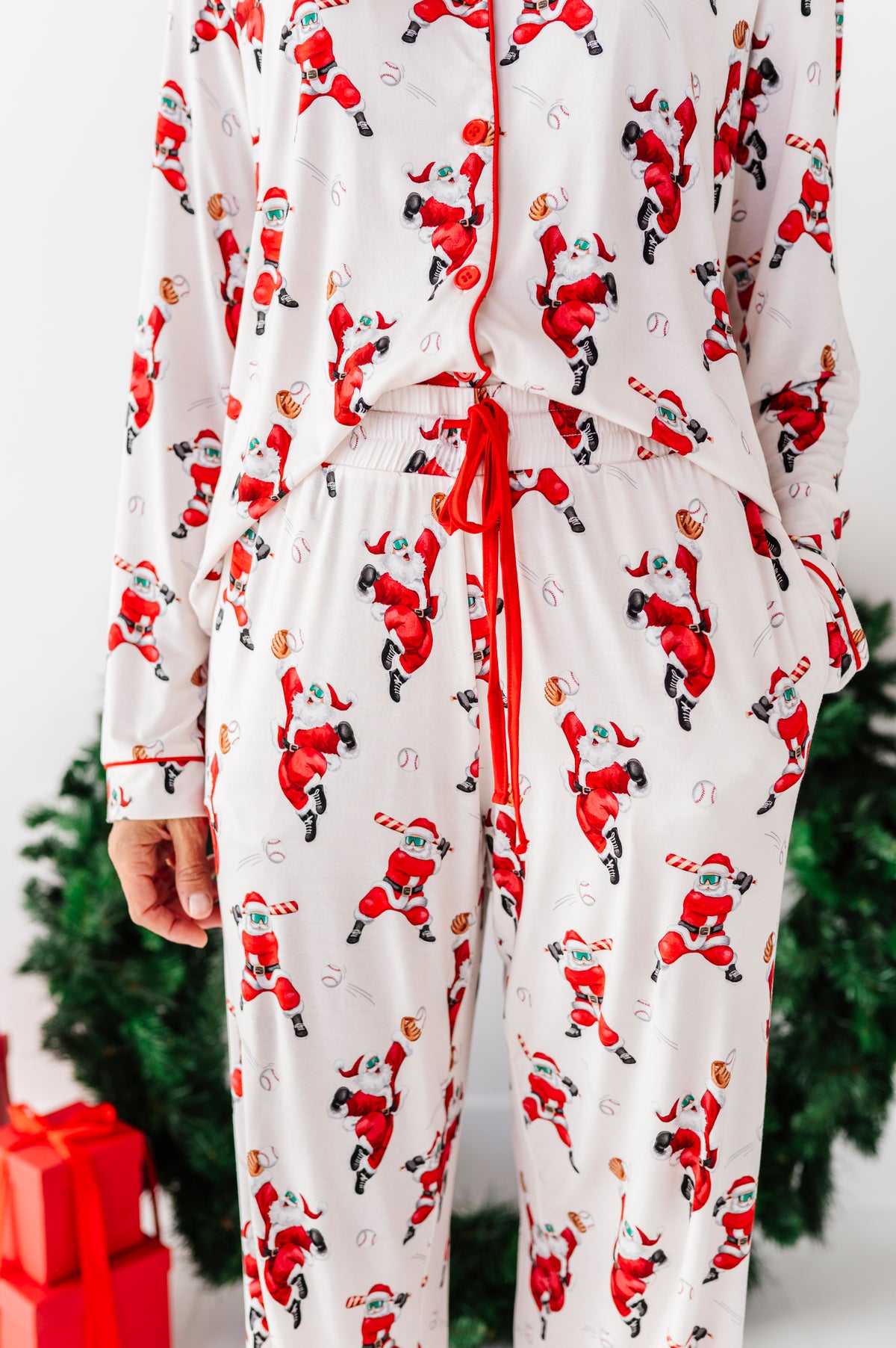 A person wearing a white long-sleeve pajama set with an allover print of Santa Claus in red and black, featuring a red drawstring waist and red piping, photographed indoors with a blurred Christmas tree and red gift boxes in the background.