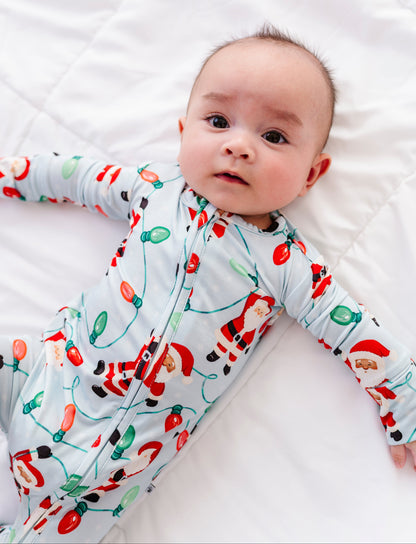 A little boy wearing a blue bamboo zip romper featuring Santa Claus hanging from multi-colored Christmas lights