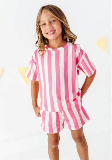 A young girl smiling and wearing a matching pink and white striped short-sleeve top and shorts set, standing against a white background with subtle yellow triangle decorations.