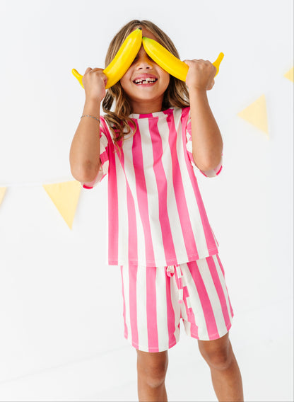A young girl wearing a pink and white vertically striped pajama set, holding two yellow bananas over her eyes in a playful pose against a white background with subtle yellow triangle decorations.