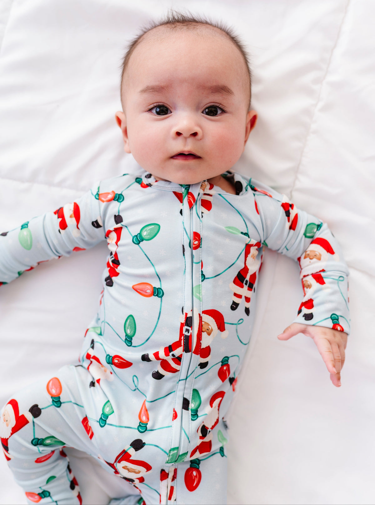 A sweet little boy wearing a blue bamboo convertible romper featuring Santa Claus hanging from multi-colored Christmas lights