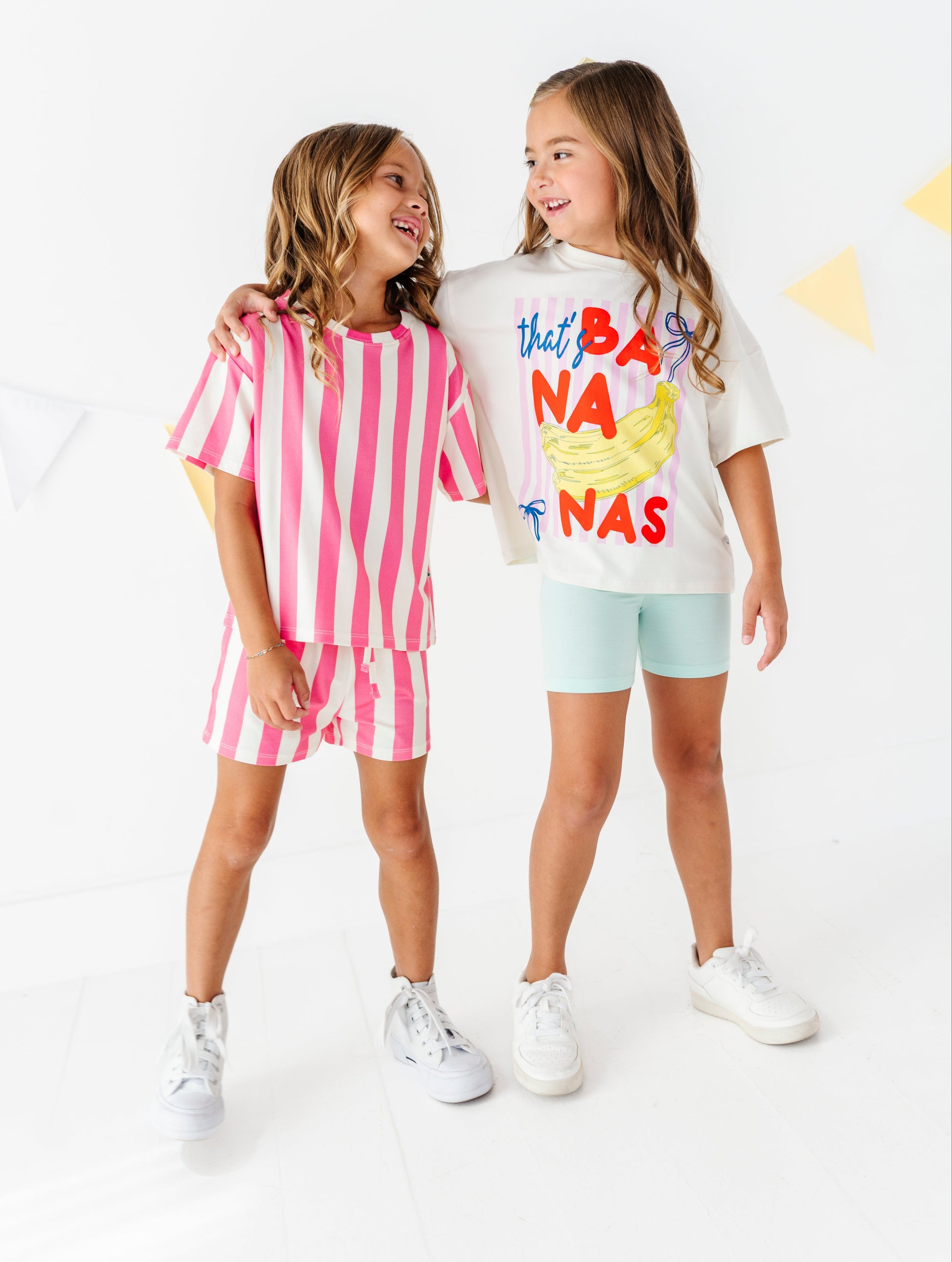 Two young girls modeling matching two-piece outfits in a bright white studio. The girl on the left wears a pink and white striped t-shirt and shorts set, while the girl on the right wears a white t-shirt with a colorful 'that's BANANA NAS' graphic and light blue shorts.