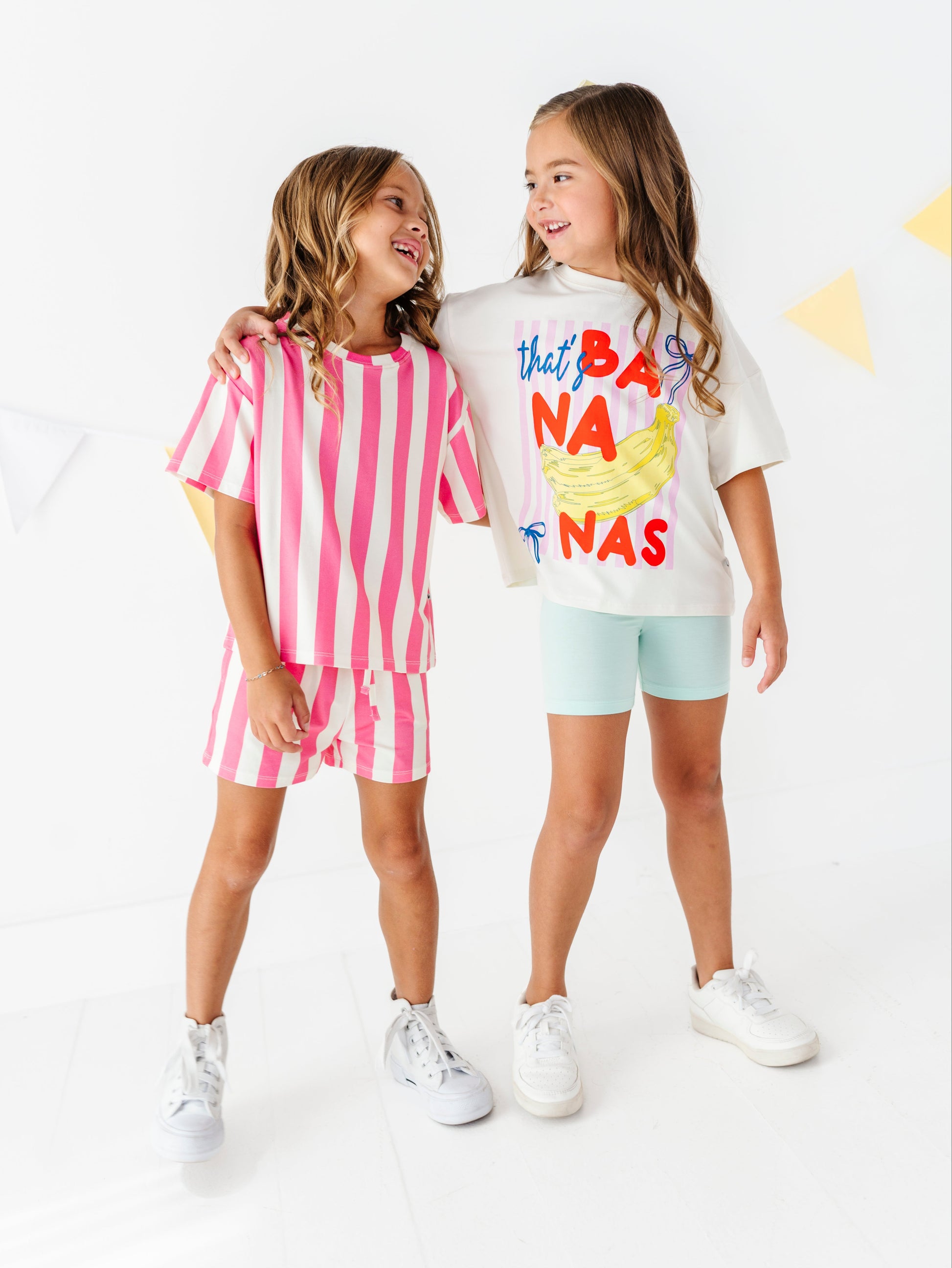 Two young girls modeling matching two-piece outfits in a bright white studio. The girl on the left wears a pink and white striped t-shirt and shorts set, while the girl on the right wears a white t-shirt with a colorful 'that's BANANA NAS' graphic and light blue shorts.