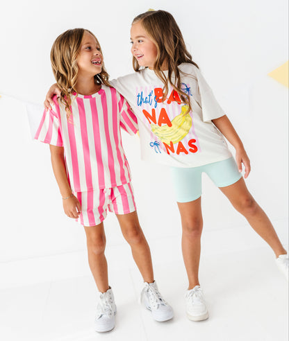 Two young girls modeling matching two-piece outfits in a bright white studio. The girl on the left wears a pink and white vertically striped t-shirt and shorts set, while the girl on the right wears a white t-shirt with a colorful banana graphic and light blue shorts.
