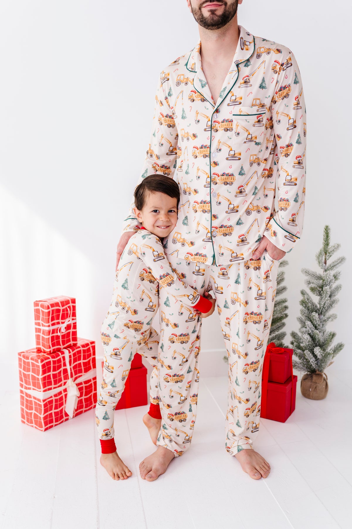 A young boy standing with his father, both wearing matching Christmas construction-themed pajamas and standing in front of a Christmas tree 