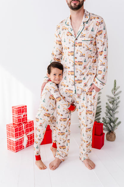 A young boy standing with his father, both wearing matching Christmas construction-themed pajamas and standing in front of a Christmas tree 