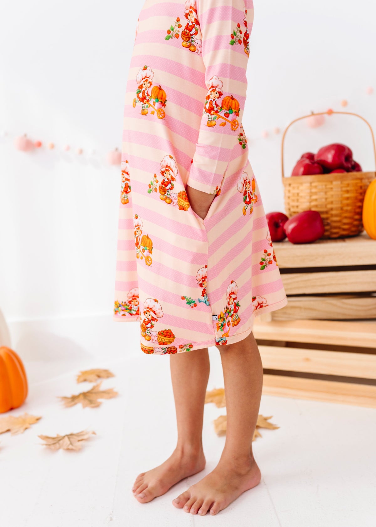 A child wearing a pink striped long-sleeve nightgown with a pumpkin and floral print, standing barefoot in a bright room with autumn decorations.
