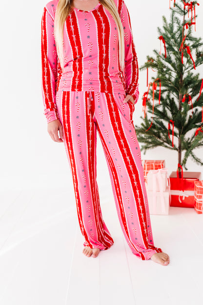 A woman wearing a festive pink and red striped long-sleeve bamboo pajama set, standing barefoot in a bright room next to a decorated Christmas tree and wrapped gifts.