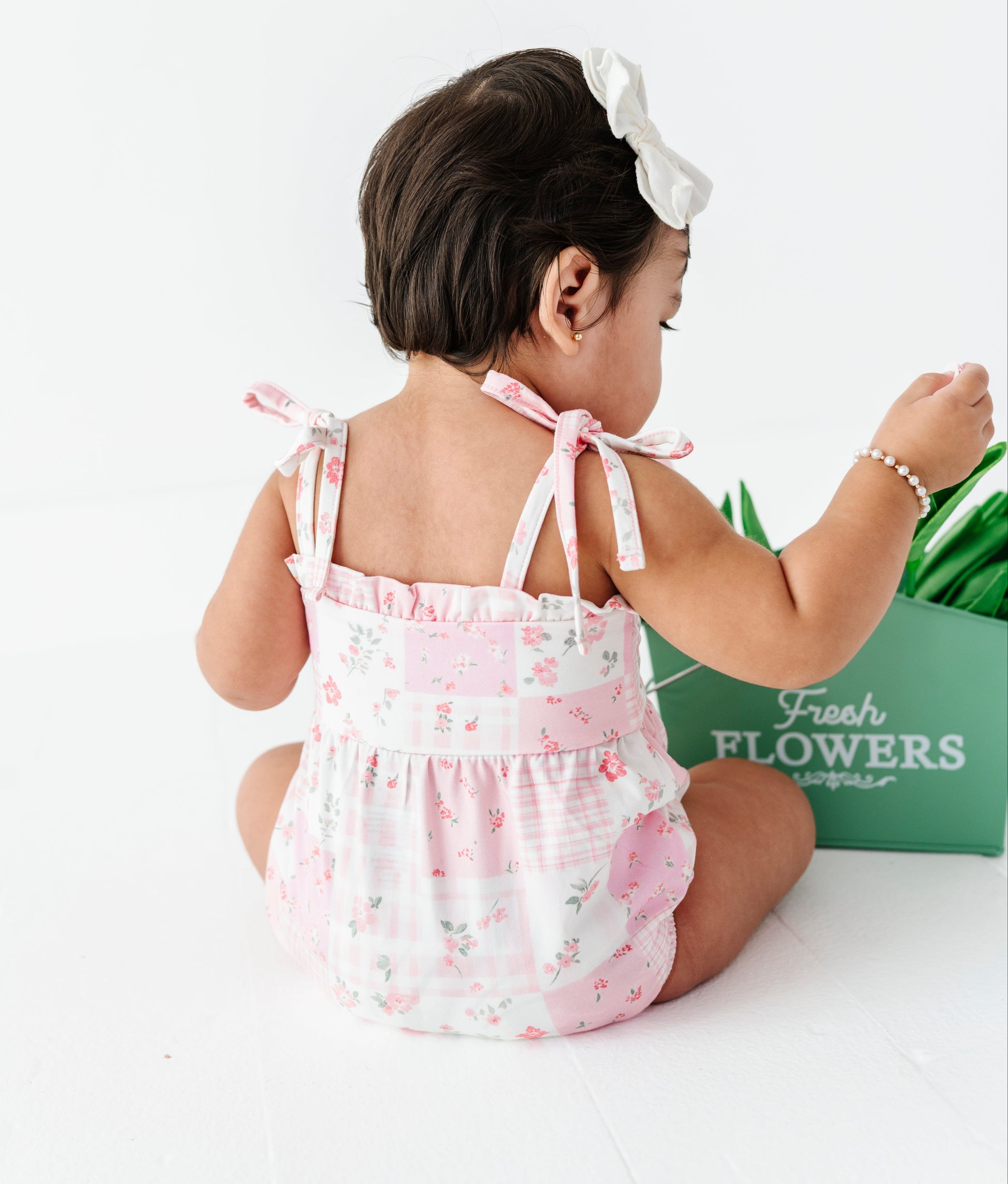 Baby in a pink floral bamboo romper sitting on a white surface with a green box labeled 'Fresh Flowers' in the background.