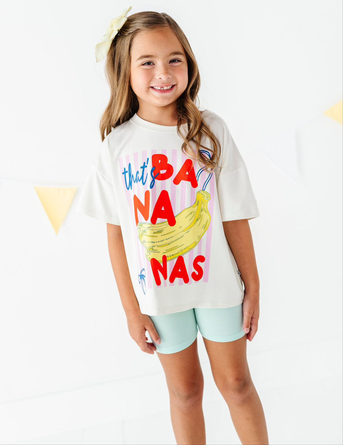 A young girl with a yellow bow in her hair stands smiling, wearing a white graphic t-shirt with a colorful 'that BANANA NAS' print and mint green shorts against a bright white background.