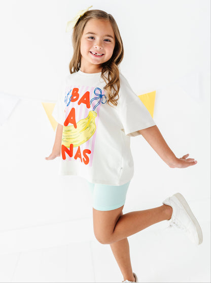 A young girl smiling and posing in a white graphic t-shirt that reads 'BANANAS' with a banana illustration, paired with light blue shorts and white sneakers against a bright white background.