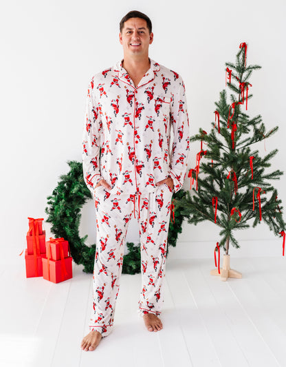 A man wearing a white long-sleeve pajama set with a red Santa Claus pattern stands barefoot in a bright white room, with a decorated Christmas tree and wrapped gifts in the background.
