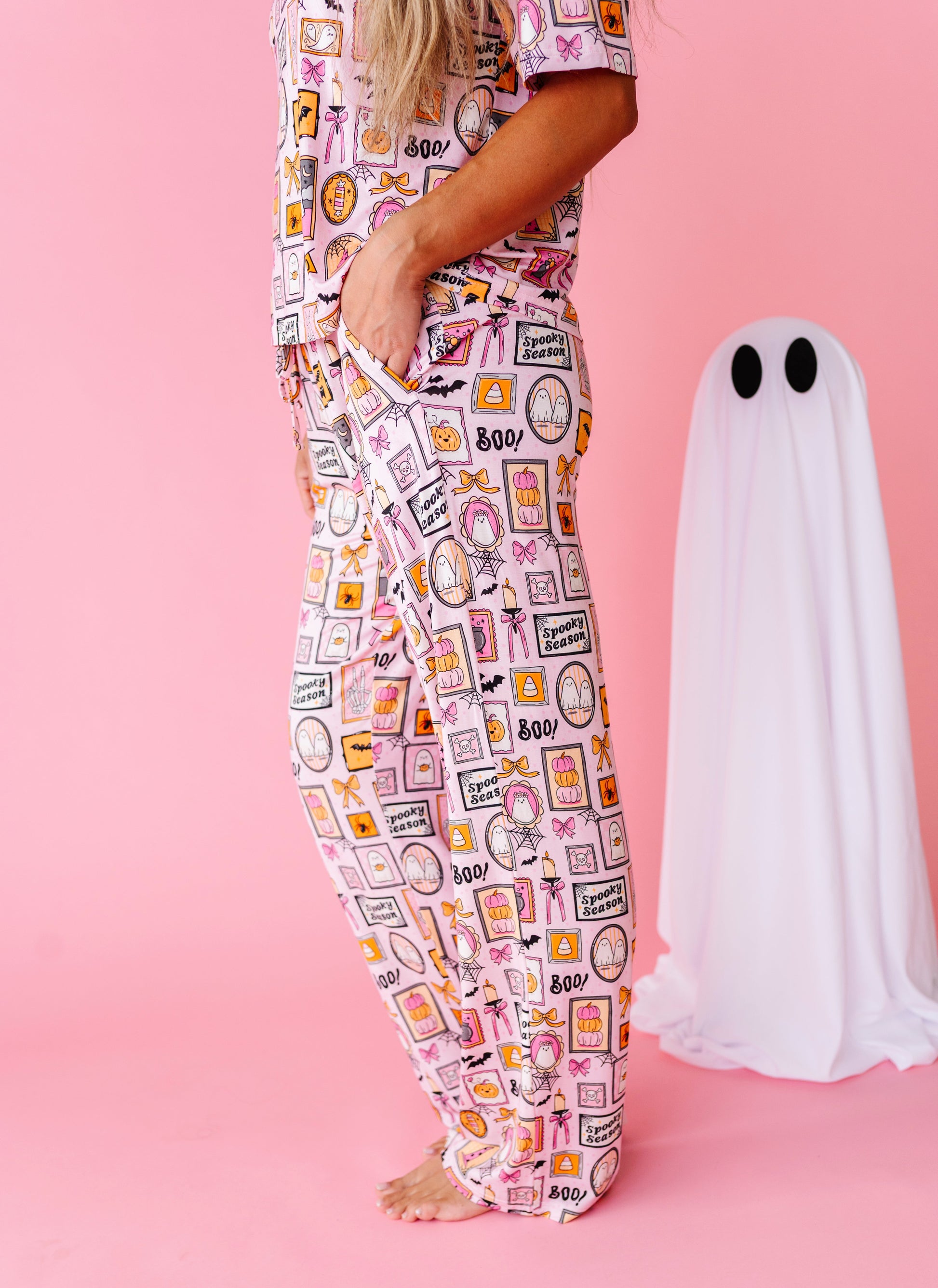 A person wearing a matching pajama set with a pink, white, and orange Halloween-themed graphic print, standing next to a white ghost decoration on a solid pink background.