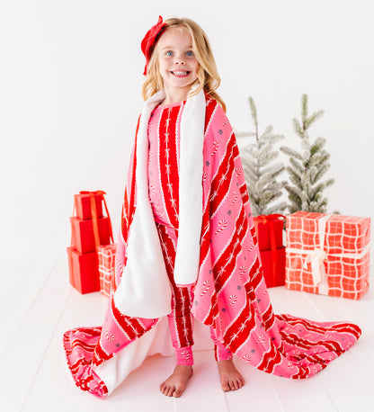 A blonde girl standing in a bright white room with red wrapped presents and a Christmas tree. She's wearing bamboo candy cane stripe pajamas and wrapped in a matching red-and-pink bamboo plush blanket 