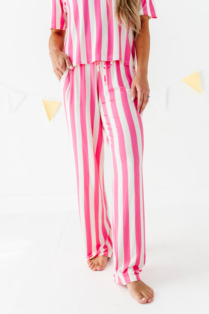 A woman wearing a pink and white vertically striped pajama set, featuring a short-sleeve top and matching pants, standing barefoot against a white background with subtle yellow bunting.