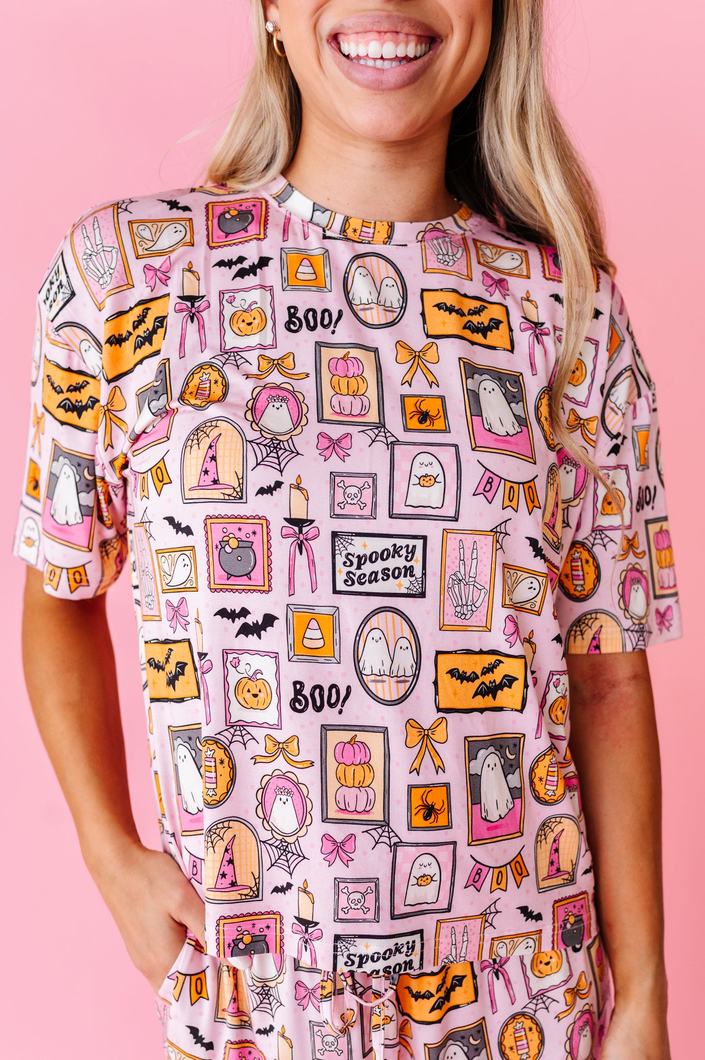 A woman wearing a pink short-sleeve t-shirt with a colorful 'Spooky Season' Halloween graphic print, set against a solid pink background.