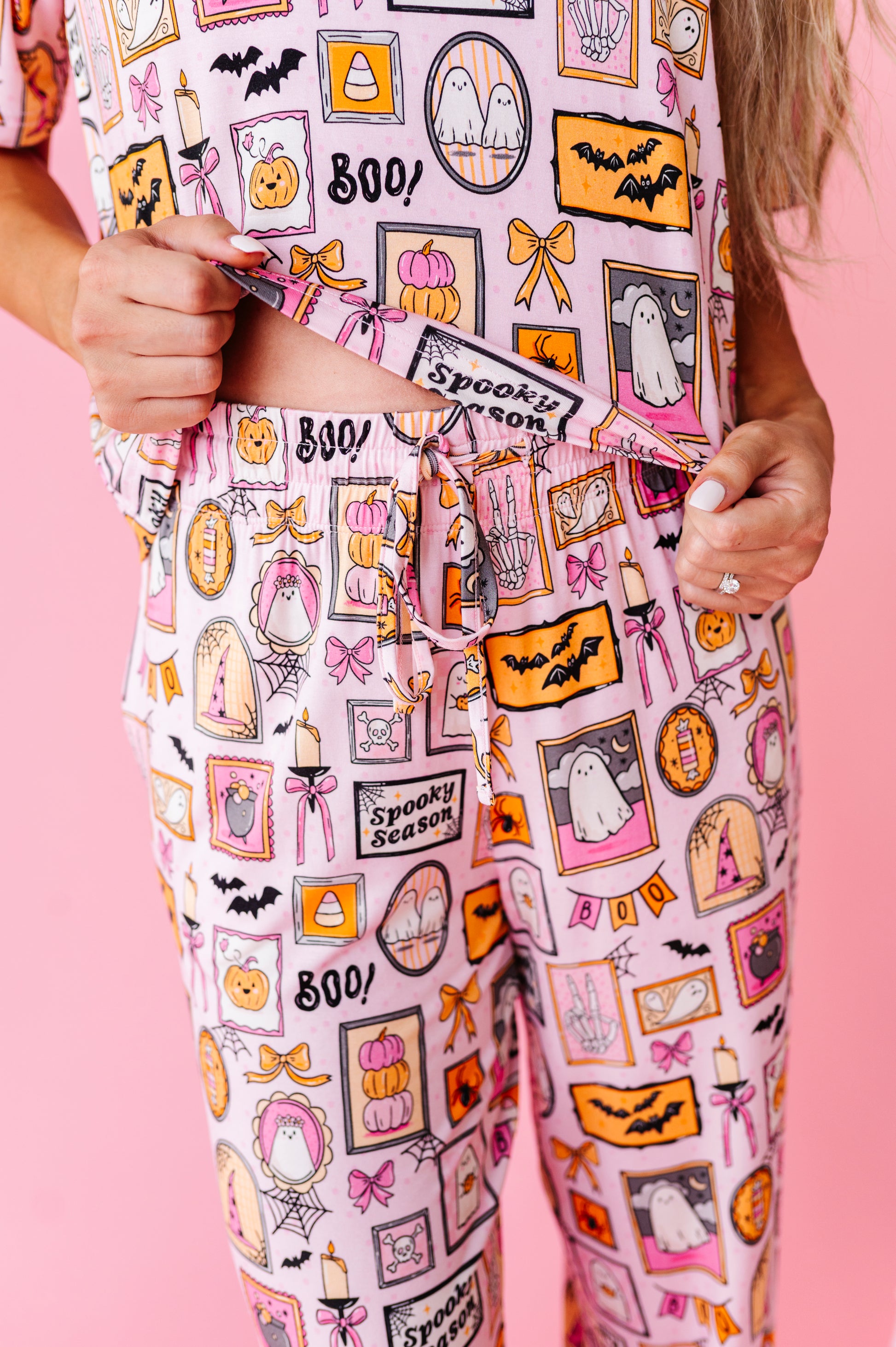 A person wearing a light pink pajama set with a colorful Halloween-themed print, including pumpkins, ghosts, bats, and 'Spooky Season' text, posed against a solid pink background.