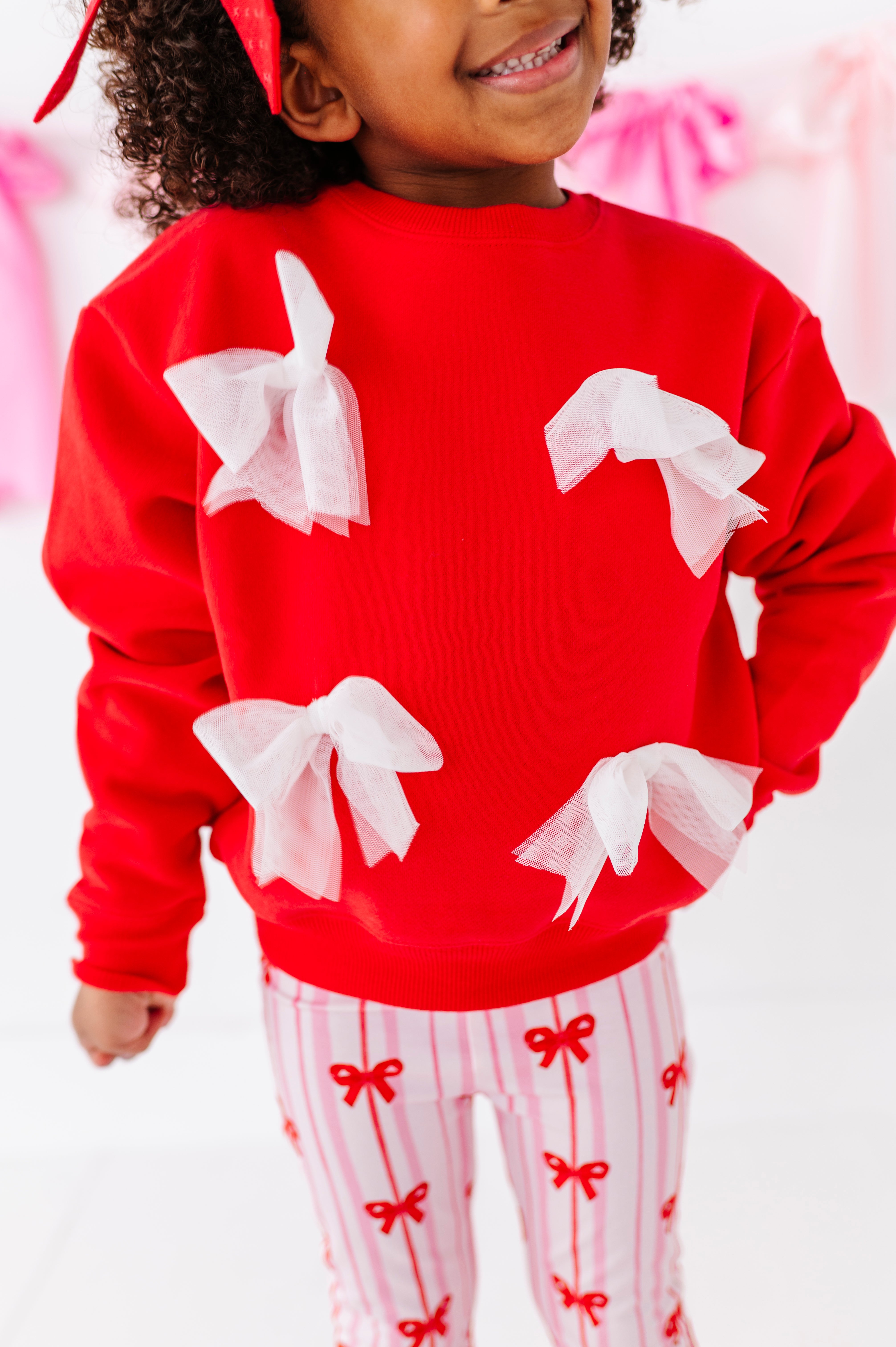 Child wearing a red crewneck sweater with white bows and striped bamboo pants with red bow patterns.