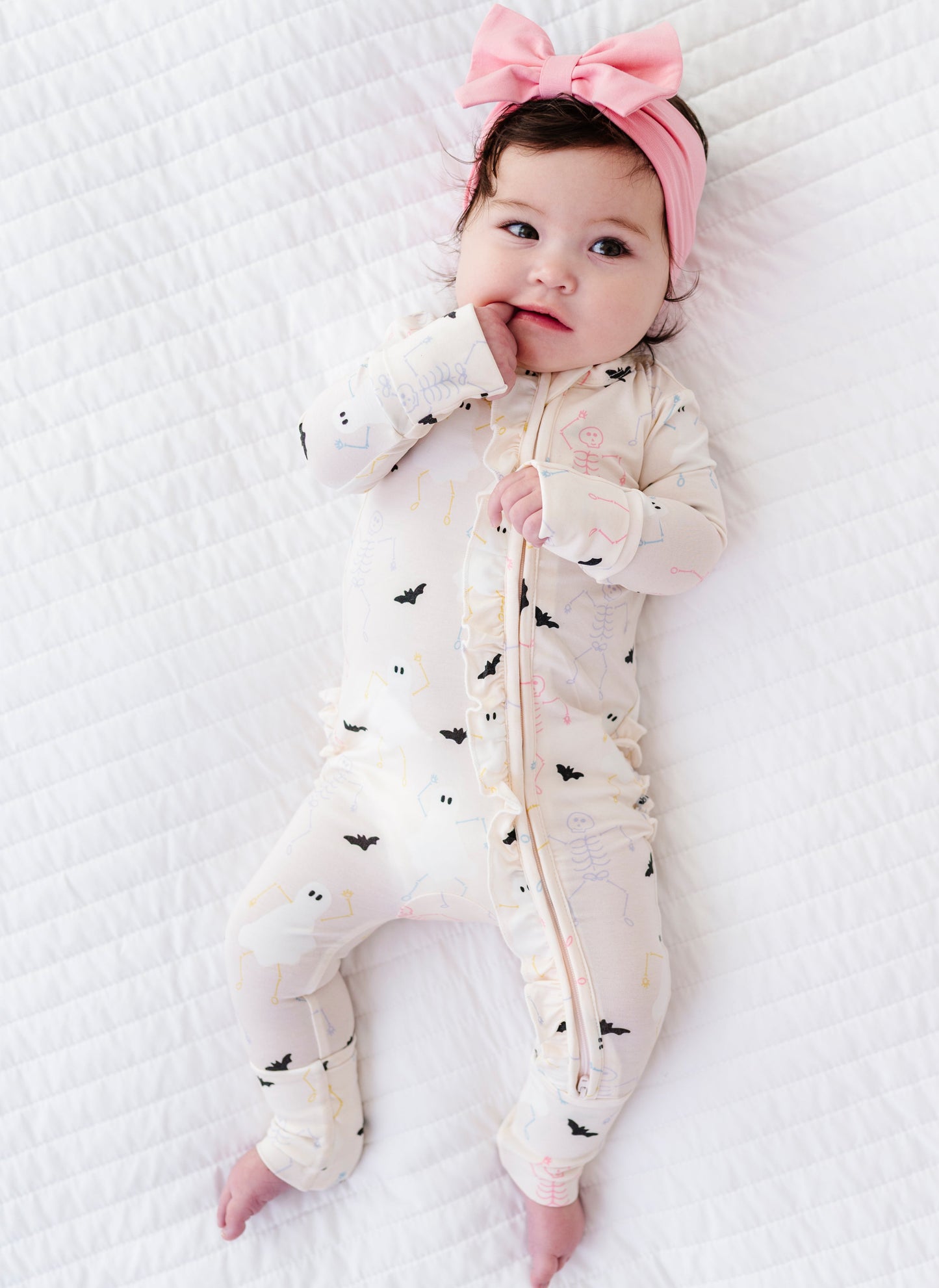 Baby lying on a white textured surface wearing a white onesie with black bat and halloween patterns and a pink headband.