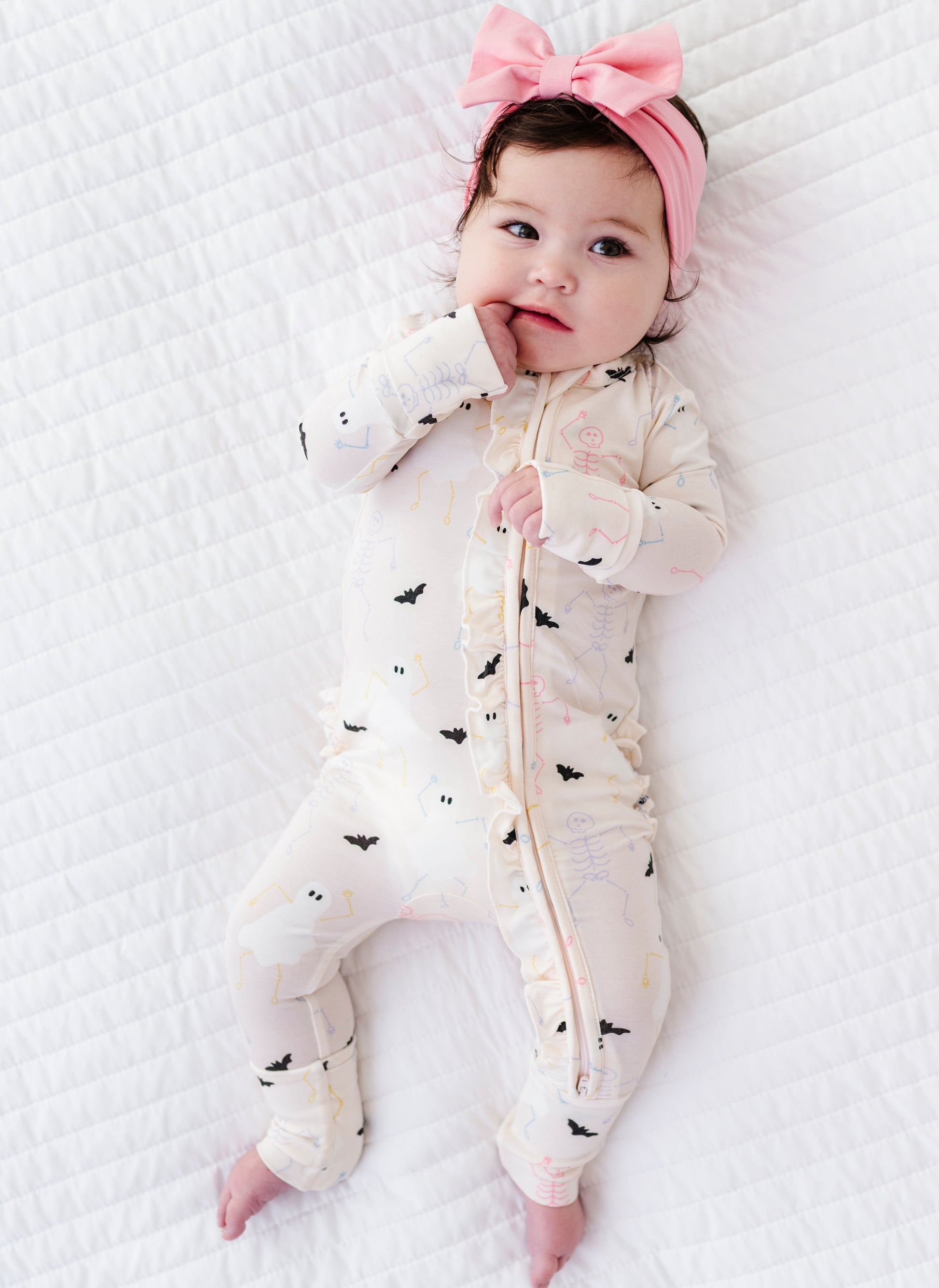 Baby lying on a white textured surface wearing a white onesie with black bat and halloween patterns and a pink headband.