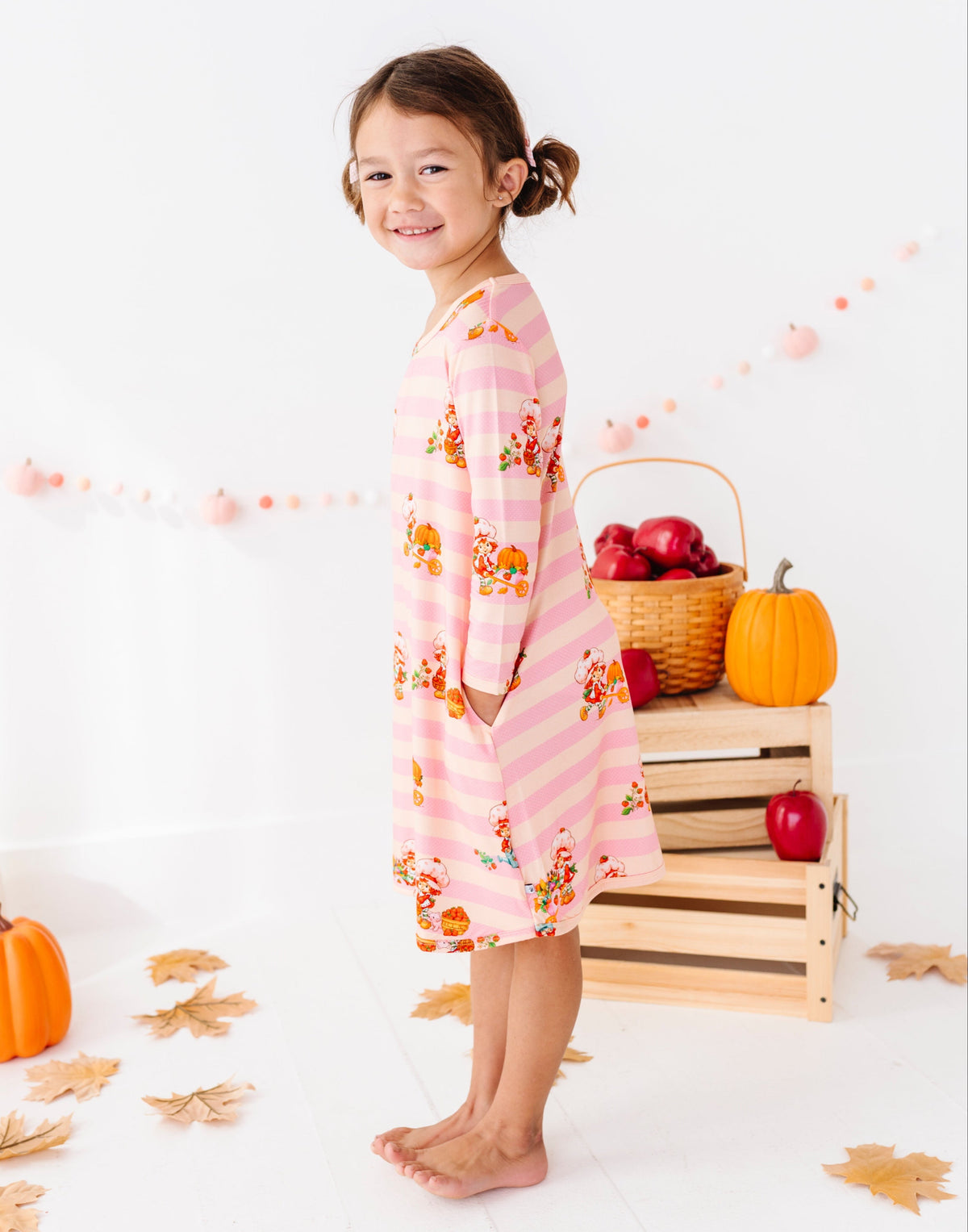 A young girl stands barefoot in a studio setting wearing a pink long-sleeve dress with horizontal stripes and a fall harvest print of pumpkins, leaves, and fruit. The background is white with decorative autumn props including pumpkins, leaves, and a basket of red apples.