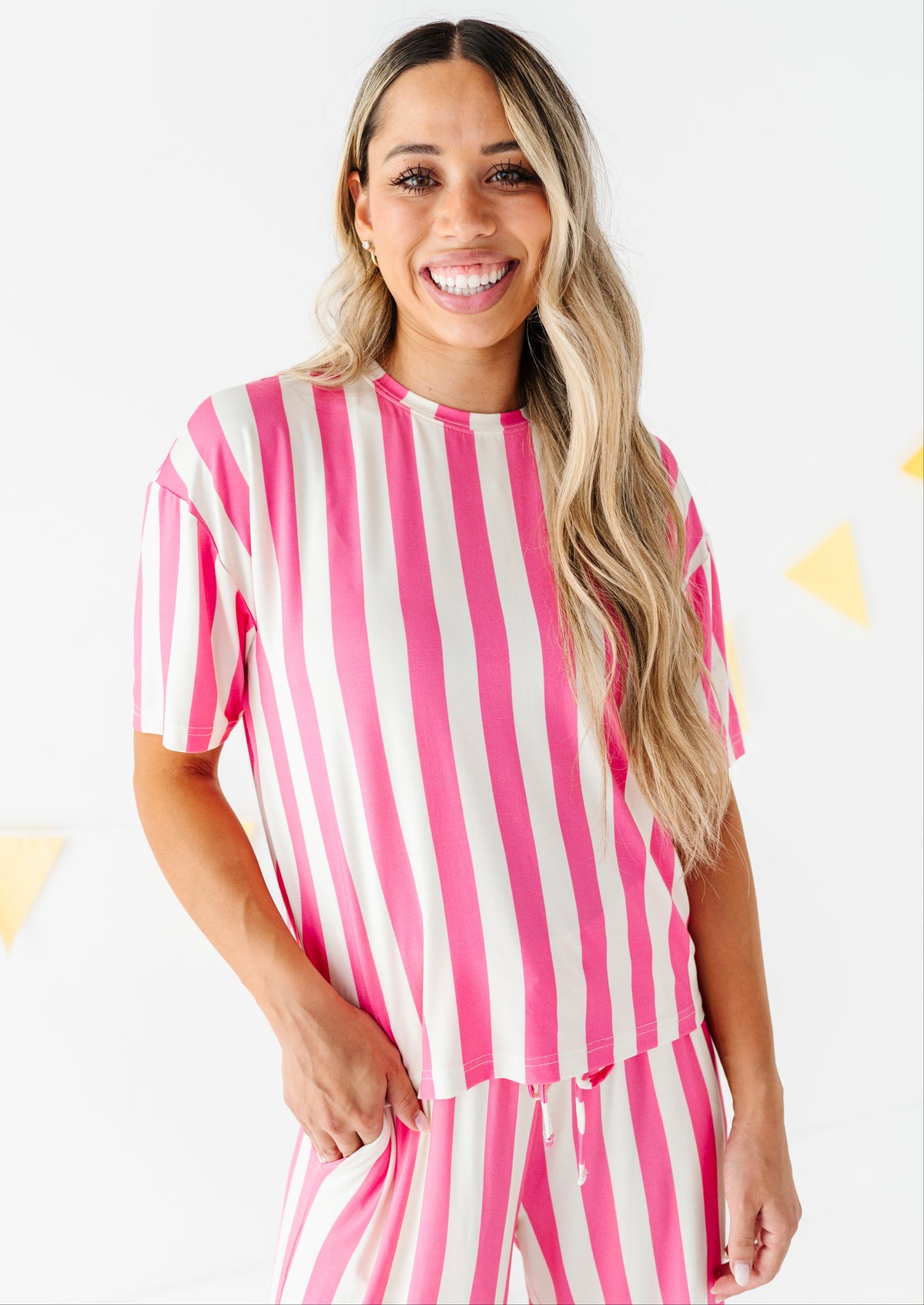 A smiling woman with long blonde-highlighted hair wearing a pink and beige vertically striped short-sleeve top and matching striped pants, standing against a white wall with small yellow triangle decorations.