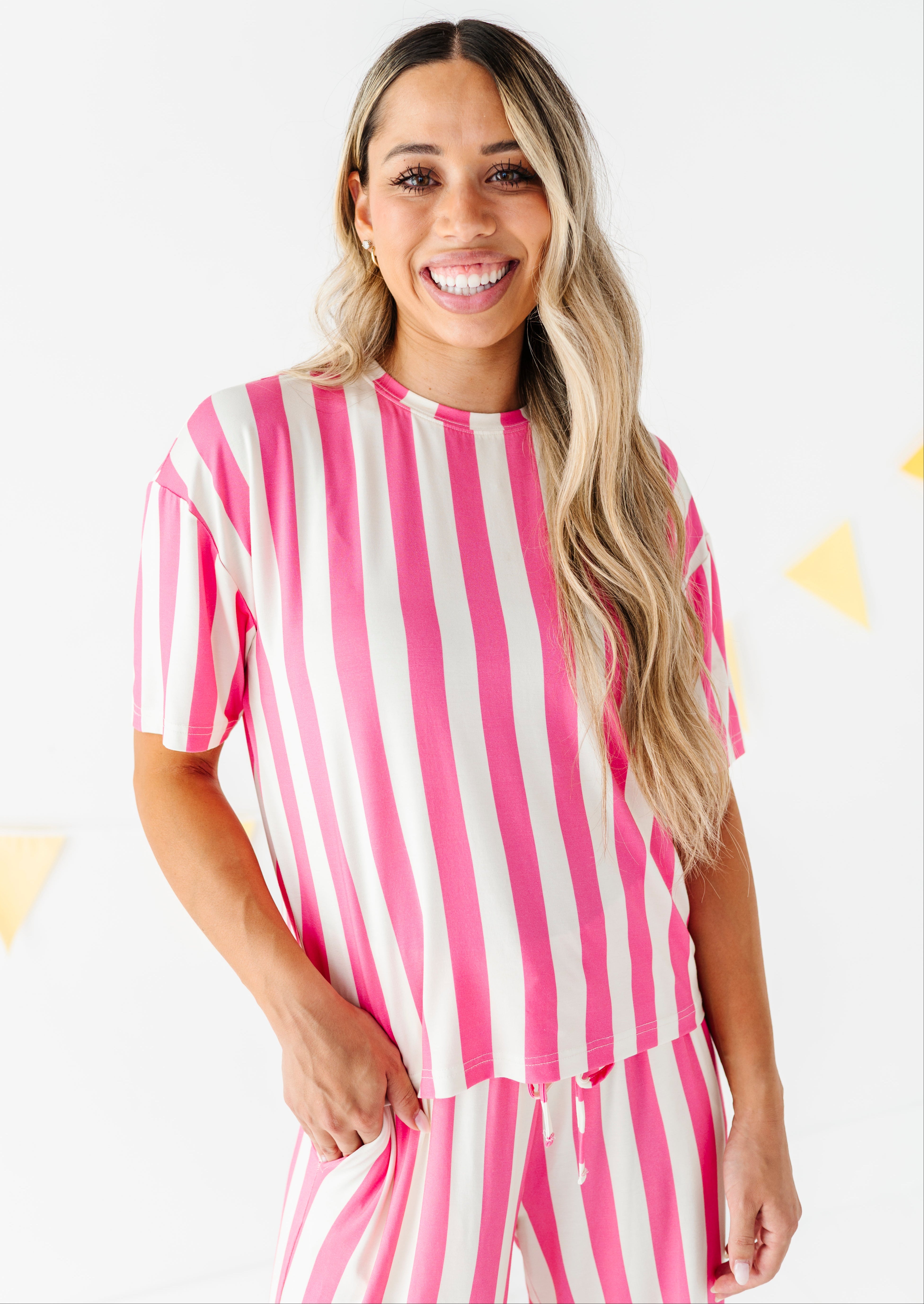 A smiling woman with long blonde-highlighted hair wearing a pink and beige vertically striped short-sleeve top and matching striped pants, standing against a white wall with small yellow triangle decorations.