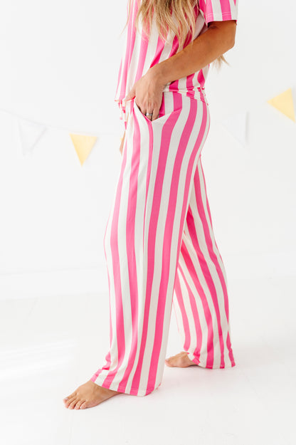 A woman wearing a pink and white vertically striped pajama set with wide-leg pants and a matching short-sleeve top, standing barefoot against a white studio background with subtle yellow bunting.