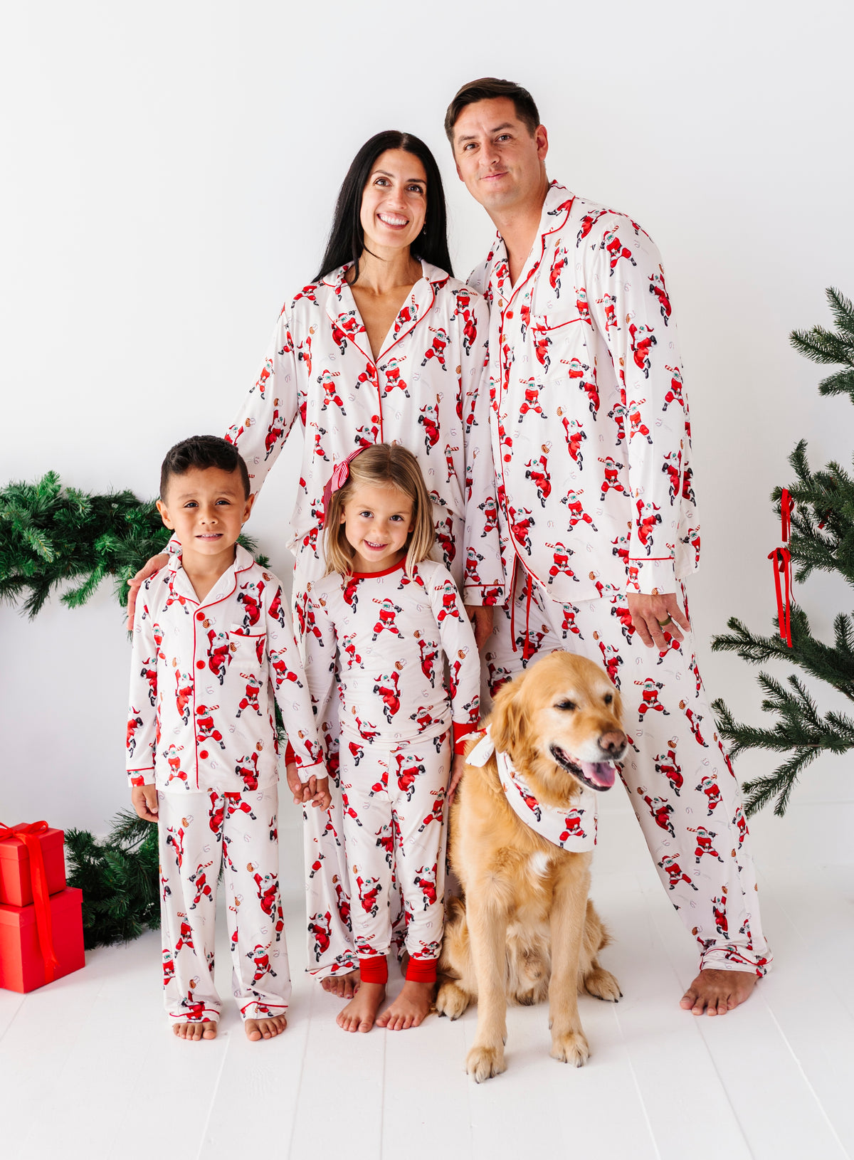A family of four wearing matching white Santa Claus patterned pajamas stands with their golden retriever dog on a white floor against a white wall, with Christmas tree branches and red gifts visible.