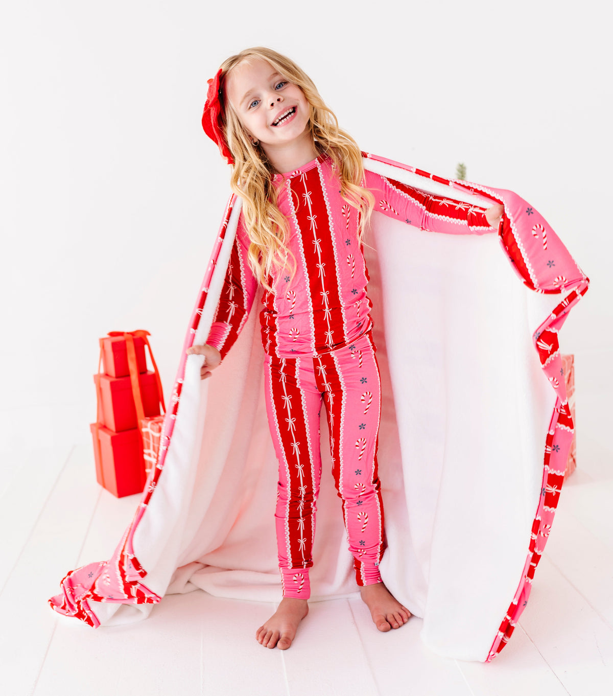 A blonde girl standing in a bright white room with red wrapped presents and a Christmas tree and smiling at the camera. She's wearing bamboo candy cane stripe pajamas and wrapped in a matching red-and-pink bamboo plush blanket 
