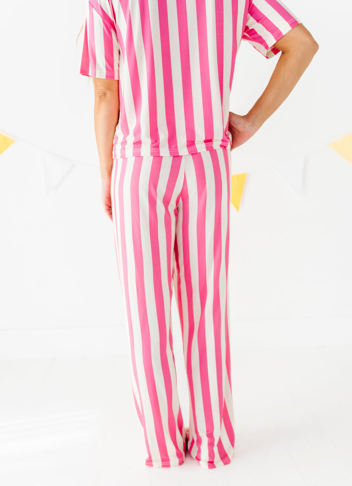 A person wearing a matching pink and white vertically striped short-sleeve top and wide-leg pants, shown from the back against a clean white studio background with faint yellow decorative shapes.