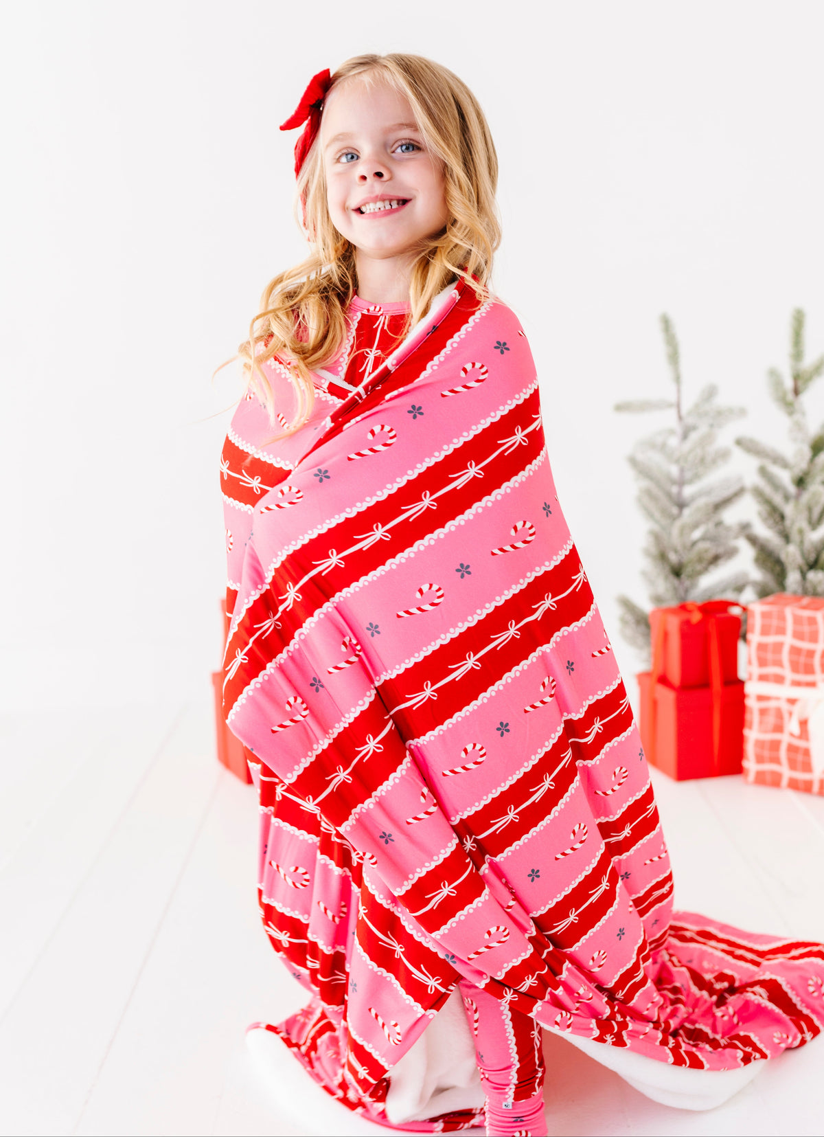 A young blonde girl standing in a bright white room with red wrapped presents and a Christmas tree. She's wearing bamboo candy cane stripe pajamas and wrapped in a matching red-and-pink bamboo plush blanket 