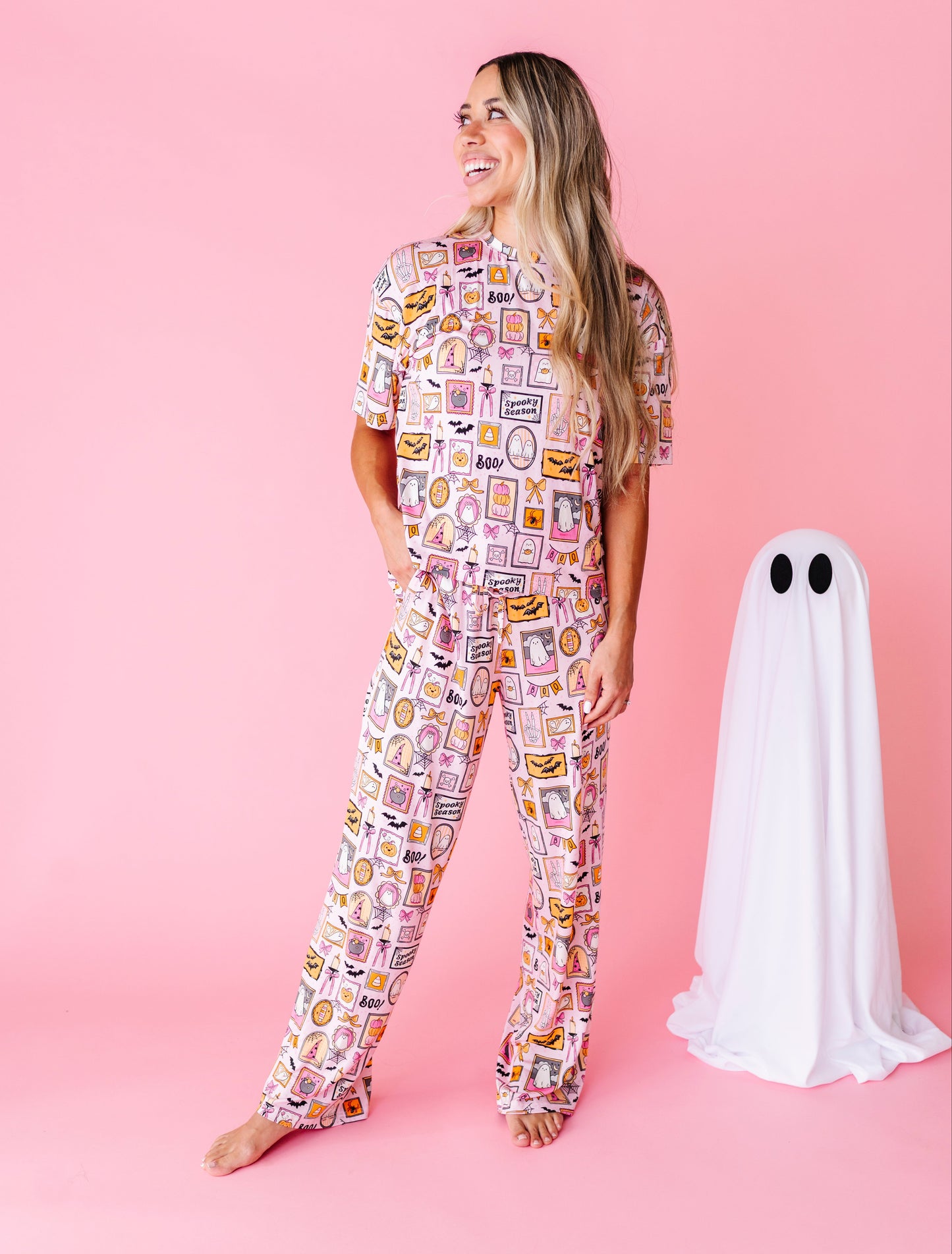 A woman stands barefoot against a pink studio background wearing a short-sleeve pajama set covered in a colorful food-themed print. To her right is a white ghost costume prop with black eyes.