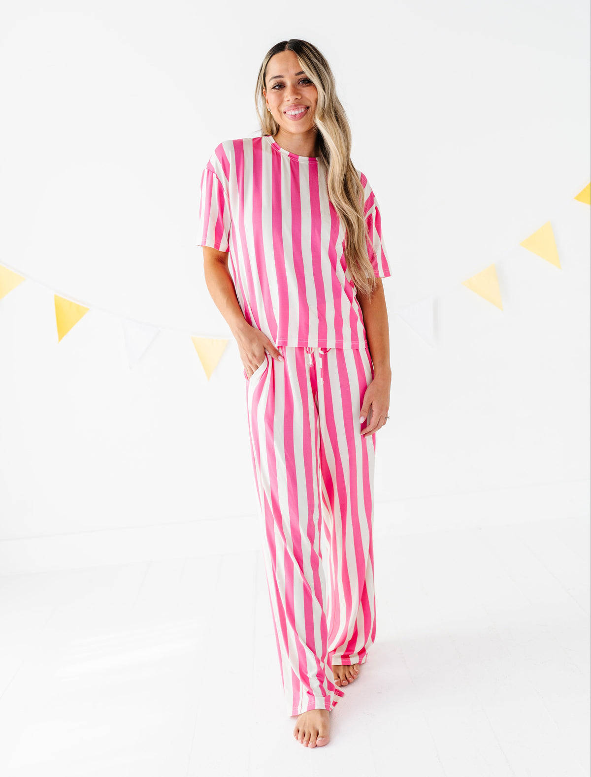 A woman modeling a pink and white vertically striped two-piece lounge set consisting of a short-sleeve top and matching pants, standing against a white studio background with yellow bunting.
