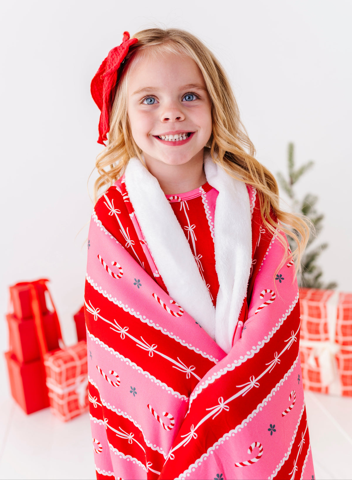 A little girl standing in a bright white room with red wrapped presents and a Christmas tree. She's wearing bamboo candy cane stripe pajamas and wrapped in a matching red-and-pink bamboo plush blanket 