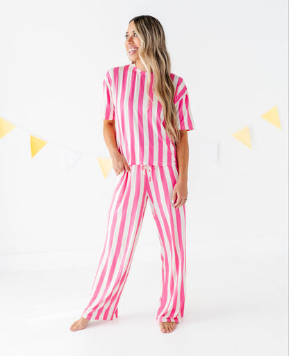 A woman modeling a matching two-piece pajama set with pink and white vertical stripes, standing against a white studio background with a string of yellow and white bunting.