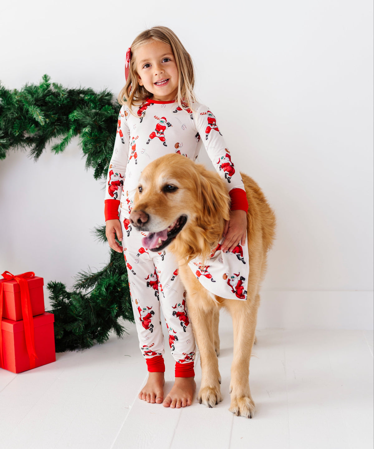 A young girl and her golden retriever dog wearing matching white Christmas-themed pajamas with a Santa Claus print, standing in front of a white background with a green wreath and red wrapped gifts.