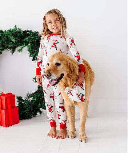 A young girl and her golden retriever dog wearing matching white Christmas-themed pajamas with a Santa Claus print, standing in front of a white background with a green wreath and red wrapped gifts.