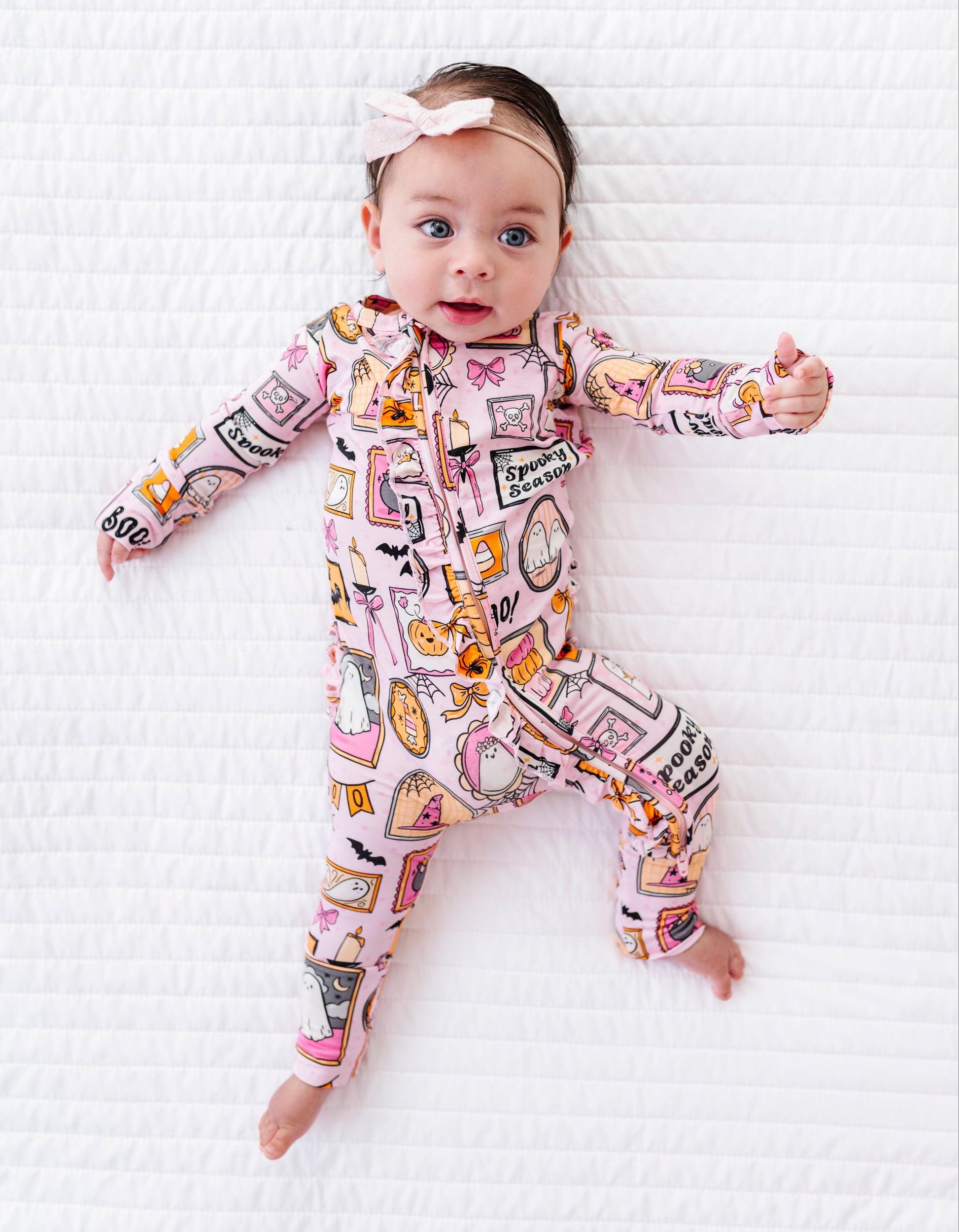 A baby lying on a white quilted blanket, wearing a pink long-sleeve footless onesie with a colorful illustrated print and a matching pink bow headband.