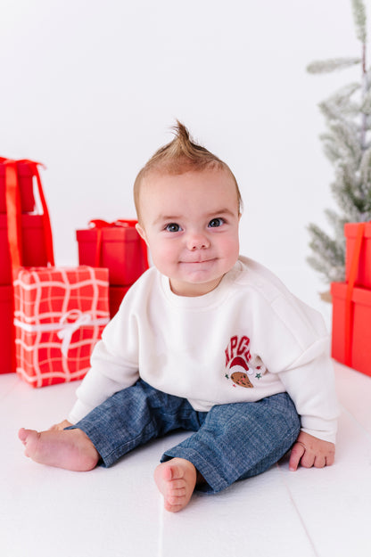 A woman and a young boy wearing matching white holiday sweatpants and sweatshirts with red and pink Christmas graphics, standing in a bright studio with wrapped presents and a Christmas garland.
